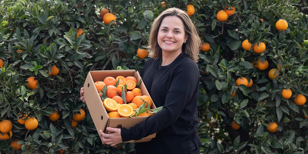 Frau mit einer Kiste Orangen in den Armen vor Orangebäumen