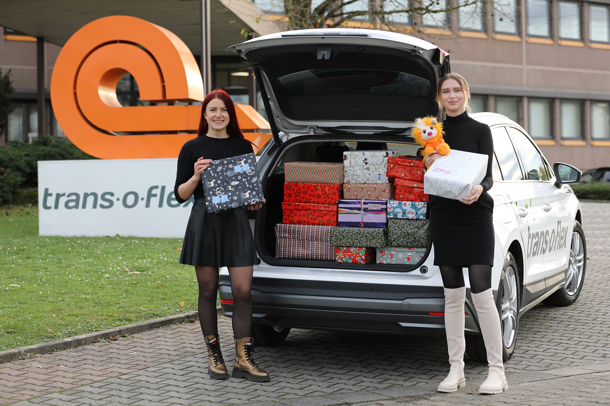 Zwei trans-o-flex-Mitarbeiterinnen stehen vor dem offenen Kofferraum eines trans-o-flex-Fahrzeugs voll mit Weihnachtspäckchen